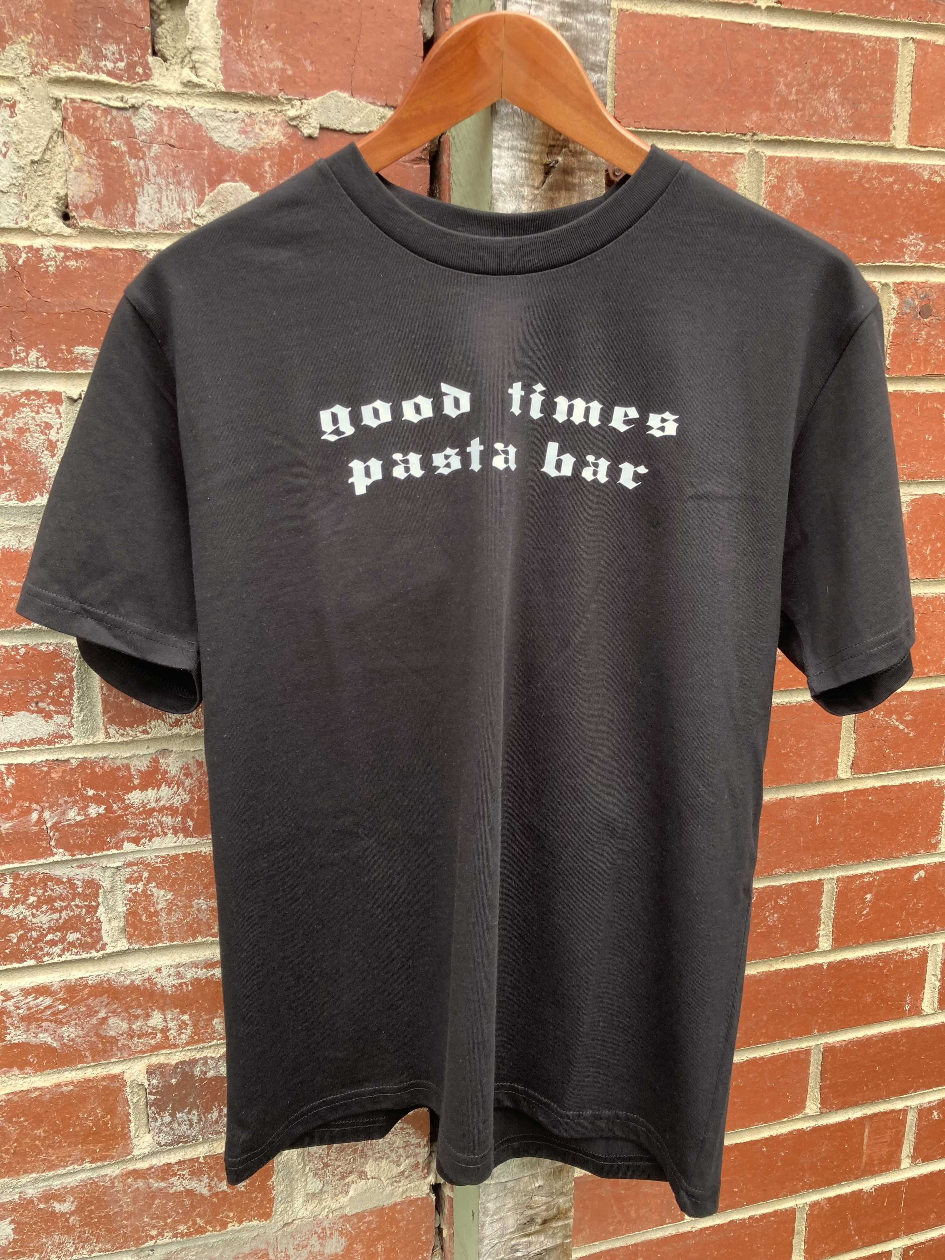 Black t-shirt with 'good times pasta bar' text on a hanger against a brick wall.