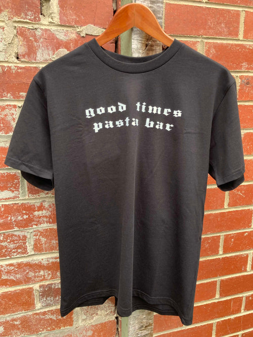 Black t-shirt with 'good times pasta bar' text on a hanger against a brick wall.